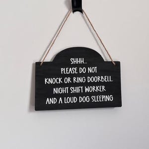 May include: A black rectangular sign with a curved top, hanging by a brown string. White text reads: "SHHH... PLEASE DO NOT KNOCK OR RING DOORBELL. NIGHT SHIFT WORKER AND A LOUD DOG SLEEPING."