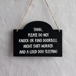 May include: A rectangular black hanging sign with a rounded top edge and a jute twine cord. The sign features white text that reads: SHHH... PLEASE DO NOT KNOCK OR RING DOORBELL. NIGHT SHIFT WORKER AND A LOUD DOG SLEEPING.