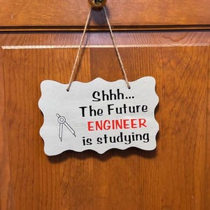 May include: A white wooden sign with a wavy edge hangs from a string. The sign says "Shhh... The Future ENGINEER is studying" in black and red lettering. A compass is drawn in black on the left side of the sign.