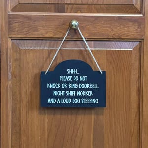 May include: A rectangular black sign with white text hanging from a jute twine cord. The text reads: SHHH... PLEASE DO NOT KNOCK OR RING DOORBELL. NIGHT SHIFT WORKER AND A LOUD DOG SLEEPING. The sign features a curved top edge and is suspended by a small knob.
