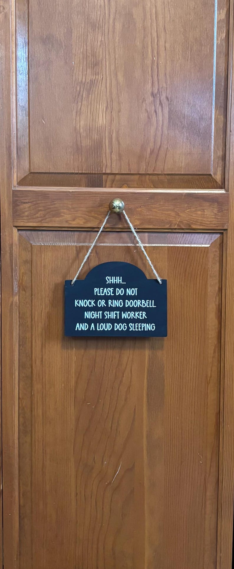 May include: A rectangular black sign with white text hanging from a jute twine cord. The text reads: SHHH... PLEASE DO NOT KNOCK OR RING DOORBELL. NIGHT SHIFT WORKER AND A LOUD DOG SLEEPING. The sign features a curved top edge and is suspended by a small knob.