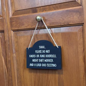 May include: A rectangular black sign with rounded top corners, featuring white text that reads: SHHH... PLEASE DO NOT KNOCK OR RING DOORBELL. NIGHT SHIFT WORKER AND A LOUD DOG SLEEPING. The sign hangs from a light-coloured twine cord.