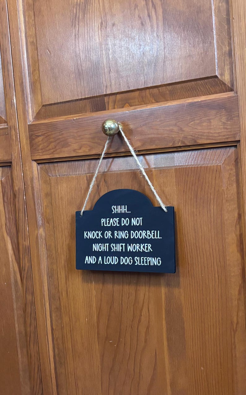 May include: A rectangular black sign with rounded top corners, featuring white text that reads: SHHH... PLEASE DO NOT KNOCK OR RING DOORBELL. NIGHT SHIFT WORKER AND A LOUD DOG SLEEPING. The sign hangs from a light-coloured twine cord.