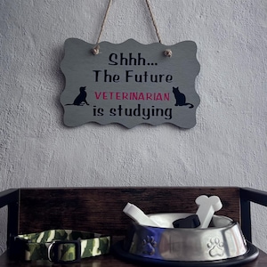 May include: A gray wooden sign with a scalloped edge hangs by a rope, featuring the black text "Shhh... The Future VETERINARIAN is studying" with black silhouettes of a dog and cat. Below, a metal bowl contains white bone-shaped toys.