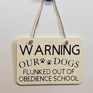 May include: A white wooden sign with black text that reads "WARNING OUR DOGS FLUNKED OUT OF OBEDIENCE SCHOOL". The sign has two paw prints between the words "OUR" and "DOGS". The sign is hanging from a string.
