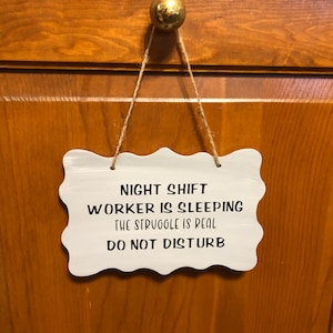 May include: White sign with rounded edges and black text that reads "Night Shift Worker is Sleeping The Struggle is Real Do Not Disturb". The sign is hanging from a wooden door with a string.