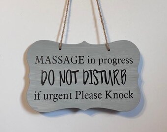 Puede incluir: Un letrero de madera gris con un borde decorativo, suspendido por un cordón de hilo natural. El letrero dice "MASSAGE in progress DO NOT DISTURB if urgent Please Knock" en letras negras, ideal para un spa o masajes.