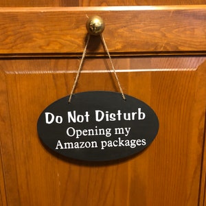 May include: A black oval sign with white text that reads "Do Not Disturb Opening my Amazon packages" hanging on a wooden door.