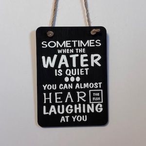 May include: Black rectangular wooden sign with white text that reads "Sometimes when the water is quiet you can almost hear the fish laughing at you." The sign is suspended by a natural fiber rope.