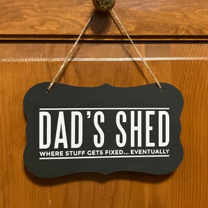 May include: Black sign with white text that reads "Dad's Shed Where Stuff Gets Fixed... Eventually". The sign is hanging from a wooden surface with twine.