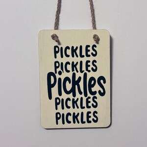 May include: A rectangular wooden sign with the word "PICKLES" repeated five times in a dark blue, bold font. The sign is cream-colored and has a natural rope hanger.