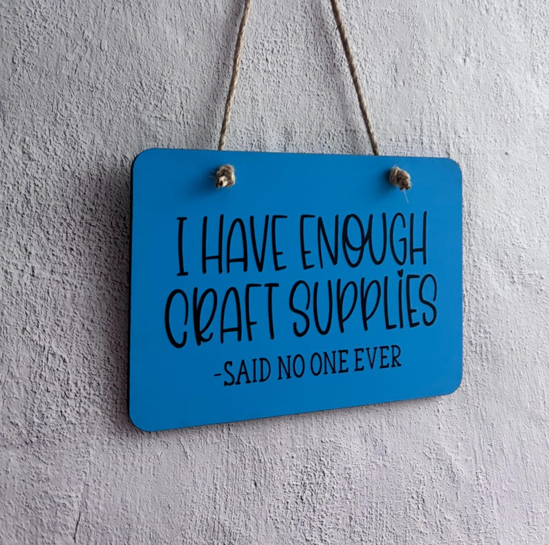 May include: A rectangular blue hanging sign with rounded corners and a jute twine cord. The sign features black text that reads: I HAVE ENOUGH CRAFT SUPPLIES -SAID NO ONE EVER. The surface of the sign is smooth and solid blue.