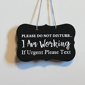 May include: Black chalkboard sign with white text that reads "Please do not disturb... I am working. If urgent please text". The sign is hanging from a string.