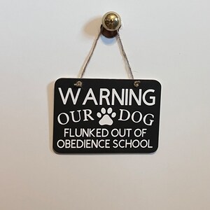 May include: Black and white sign with the text "WARNING OUR DOG FLUNKED OUT OF OBEDIENCE SCHOOL" with a paw print in the middle of the text. The sign is hanging from a string with a gold knob.