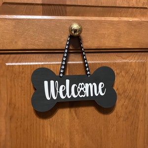 May include: Black wooden bone-shaped welcome sign with white lettering that says "Welcome" with a paw print in the "O". The sign is hanging from a black ribbon with white paw print designs.