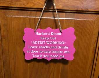 May include: A pink sign with rounded edges hangs on a wooden door. The sign reads "Harlow's Room Keep Out *ARTIST WORKING* Leave snacks and drinks at door to help inspire me. Text if you need me."