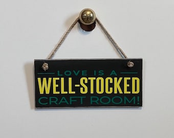 May include: Black wooden sign with white rope hanging from a silver hook. The sign reads "Love is a well-stocked craft room!" in green and yellow lettering.