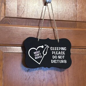 May include: Black, heart-shaped sign with white text and a stethoscope design. The sign reads "NIGHT SHIFT NURSE" inside the heart and "SLEEPING PLEASE DO NOT DISTURB" on the right. The sign hangs from a white hook.