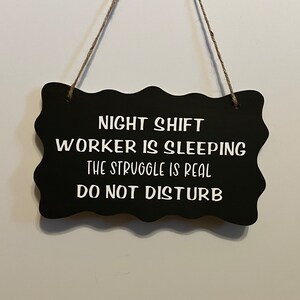 May include: Black sign with white text that reads "Night Shift Worker is Sleeping The Struggle is Real Do Not Disturb".