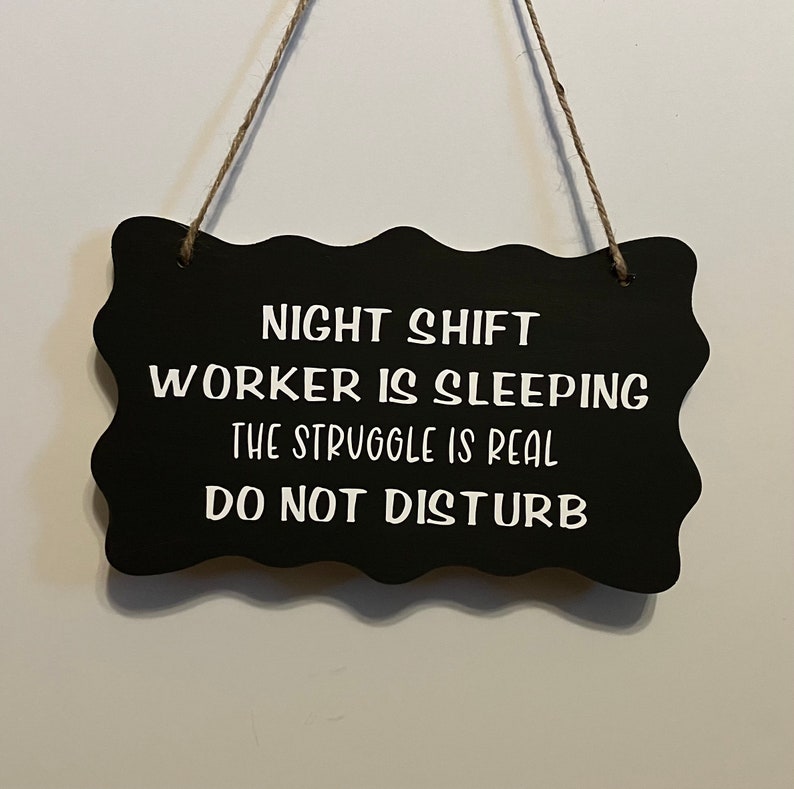 May include: Black sign with white text that reads "Night Shift Worker is Sleeping The Struggle is Real Do Not Disturb".