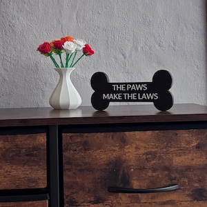 May include: A white vase with a fluted design holds a bouquet of colorful roses. A black, bone-shaped sign with the text "THE PAWS MAKE THE LAWS" stands nearby. The arrangement sits on a dark brown wooden surface.