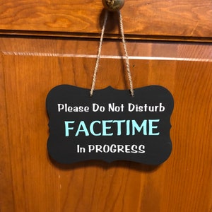 May include: Black chalkboard sign with white text that reads "Please Do Not Disturb Facetime In Progress". The sign is hanging from a wooden door with twine.
