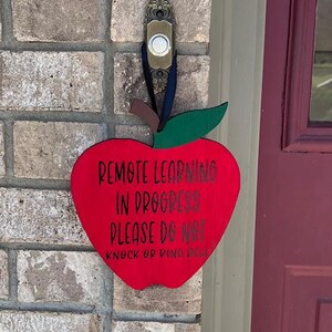 May include: A red apple-shaped sign with the text "Remote Learning in Progress Please Do Not Knock or Ring Bell" hanging on a brick wall.