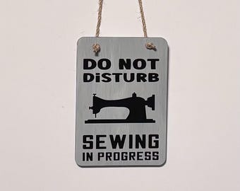 May include: A rectangular sign with the text "DO NOT DISTURB" above a black sewing machine silhouette and "SEWING IN PROGRESS" below. The sign is gray with black text and a brown string for hanging.