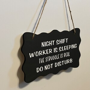 May include: Black sign with white text that reads "Night Shift Worker is Sleeping, The Struggle is Real, Do Not Disturb".