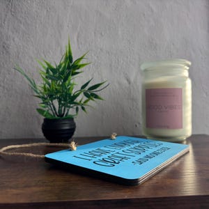 May include: A rectangular light blue wooden sign with black text that reads I HAVE ENOUGH CRAFT SUPPLIES -SAID NO ONE EVER, featuring a jute twine hanger. Next to it is a small green plant in a black pot and a white candle in a glass jar.