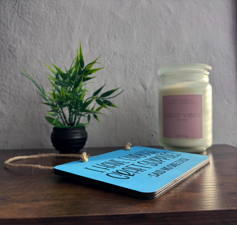 May include: A rectangular light blue wooden sign with black text that reads I HAVE ENOUGH CRAFT SUPPLIES -SAID NO ONE EVER, featuring a jute twine hanger. Next to it is a small green plant in a black pot and a white candle in a glass jar.