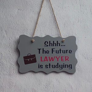 May include: A gray rectangular hanging sign with scalloped edges and a jute twine hanger. The sign features a dark brown briefcase icon and the black and pink text: Shhh... The Future LAWYER is studying, printed in a clear, sans-serif font.