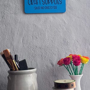 May include: A blue rectangular wall sign with the text I HAVE ENOUGH CRAFT SUPPLIES -SAID NO ONE EVER, a gray ceramic jar holding paint brushes, a white vase with colorful fabric roses, and a stack of four decorative ribbon spools.