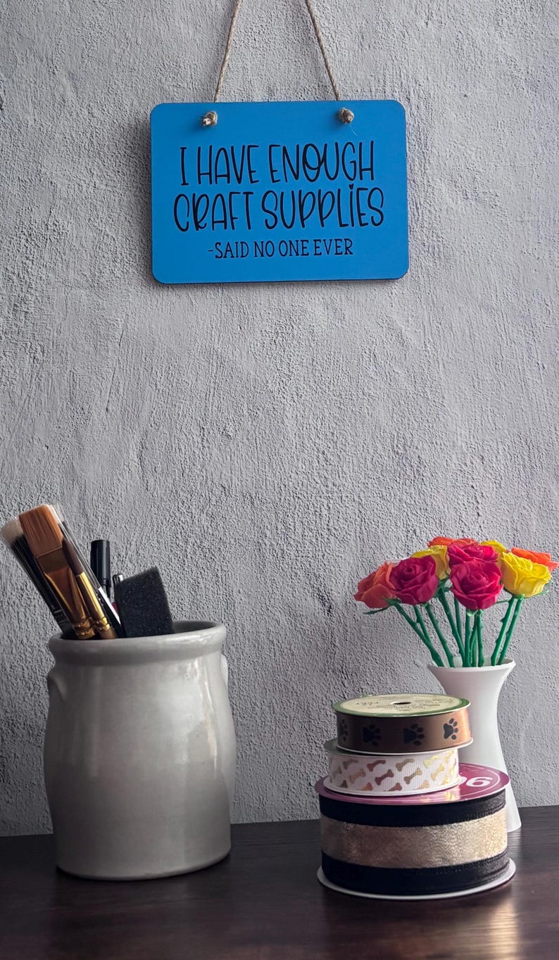 May include: A blue rectangular wall sign with the text I HAVE ENOUGH CRAFT SUPPLIES -SAID NO ONE EVER, a gray ceramic jar holding paint brushes, a white vase with colorful fabric roses, and a stack of four decorative ribbon spools.