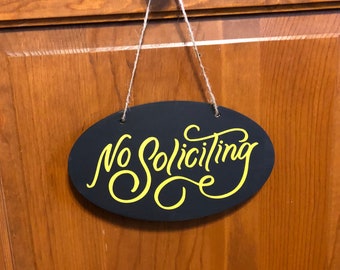 May include: A black oval sign with the text "No Soliciting" written in yellow cursive lettering. The sign is hanging from a wooden door.