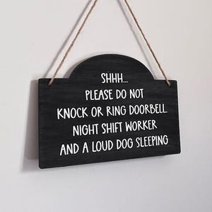 May include: A black rectangular wooden sign with a rounded top edge, suspended by a thin brown twine string. The sign features white text that reads: SHHH... PLEASE DO NOT KNOCK OR RING DOORBELL. NIGHT SHIFT WORKER AND A LOUD DOG SLEEPING.