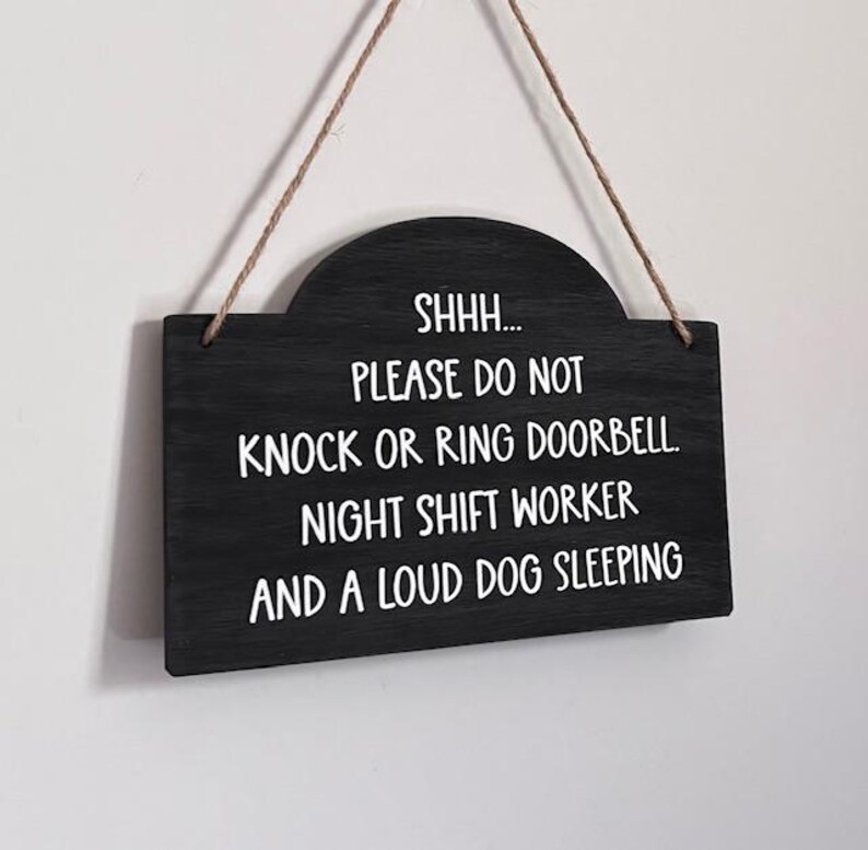 May include: A black rectangular wooden sign with a rounded top edge, suspended by a thin brown twine string. The sign features white text that reads: SHHH... PLEASE DO NOT KNOCK OR RING DOORBELL. NIGHT SHIFT WORKER AND A LOUD DOG SLEEPING.