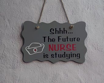 May include: A gray rectangular hanging sign with scalloped edges and a jute rope hanger. The sign features the black text "Shhh... The Future" and "is studying" with the red word "NURSE" in the center, accompanied by a white nurse cap icon.