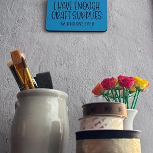 May include: A blue rectangular wall sign with the black text I HAVE ENOUGH CRAFT SUPPLIES -SAID NO ONE EVER, a gray ceramic holder with paintbrushes, and a stack of three decorative ribbons next to a small vase of colorful artificial roses.