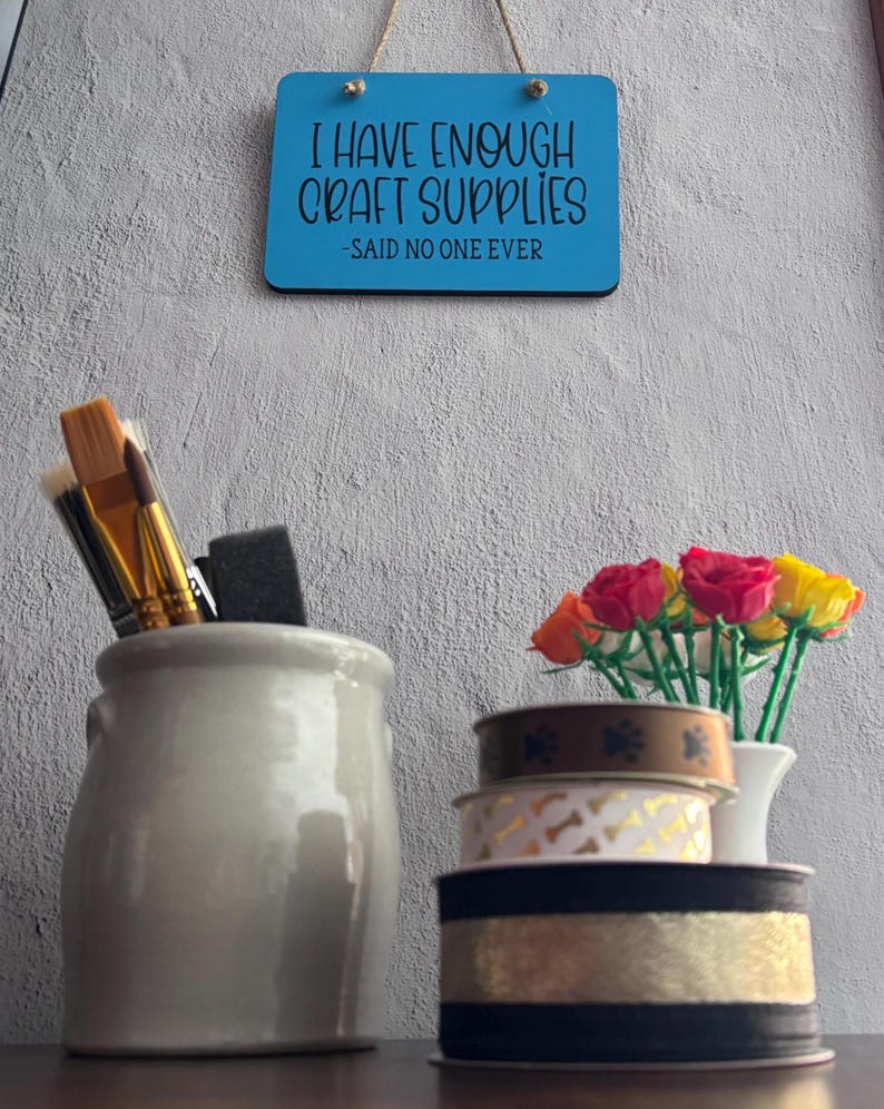 May include: A blue rectangular wall sign with the black text I HAVE ENOUGH CRAFT SUPPLIES -SAID NO ONE EVER, a gray ceramic holder with paintbrushes, and a stack of three decorative ribbons next to a small vase of colorful artificial roses.