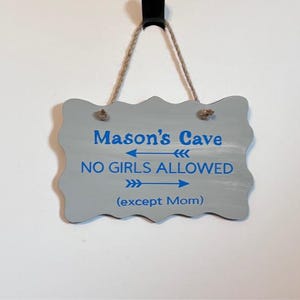 May include: A gray wooden sign with a wavy edge, hanging by a jute rope. The sign reads "Mason's Cave" above "NO GIRLS ALLOWED" in blue text, with "(except Mom)" below. The sign is hanging against a white wall.