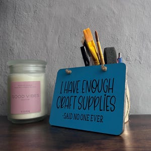 May include: A frosted glass candle jar with a pink label reading GOOD VIBES SOOTHE and a blue rectangular wooden sign with the text I HAVE ENOUGH CRAFT SUPPLIES -SAID NO ONE EVER, holding various paintbrushes and pens.
