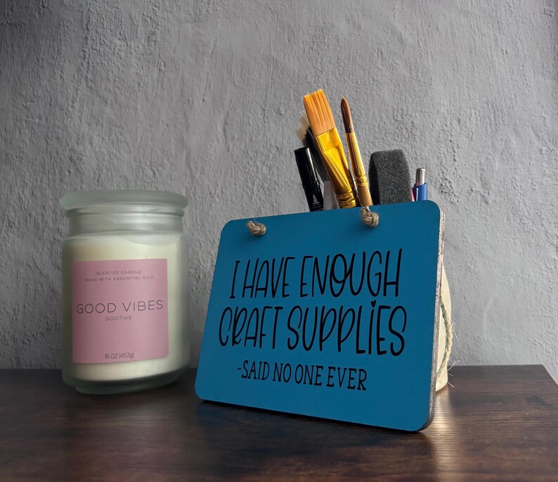 May include: A frosted glass candle jar with a pink label reading GOOD VIBES SOOTHE and a blue rectangular wooden sign with the text I HAVE ENOUGH CRAFT SUPPLIES -SAID NO ONE EVER, holding various paintbrushes and pens.