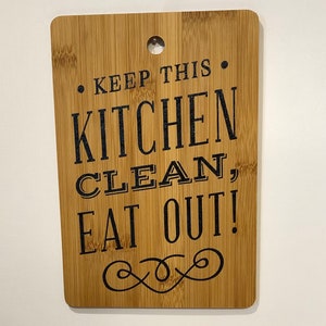 May include: A brown wooden cutting board with the text "Keep this kitchen clean, eat out!" in black lettering. The board has a decorative swirl design at the bottom.
