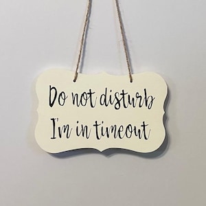 May include: A white wooden sign with a black cursive inscription that reads "Do not disturb I'm in timeout". The sign has a decorative border and is hanging from a string.
