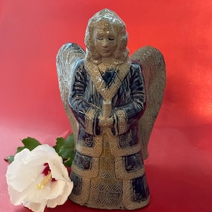 May include: A ceramic angel figurine featuring a glossy dark blue robe with textured tan trim and large wings. The figure has a tan face and hair, and holds a small cylindrical object in its hands. A white hibiscus flower is positioned nearby.