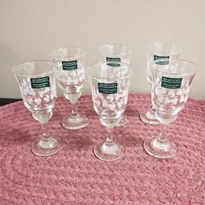 May include: Six clear glass shot glasses with a white shamrock design and the word "eamon" hand engraved in green lettering. The glasses are on a pink woven surface.