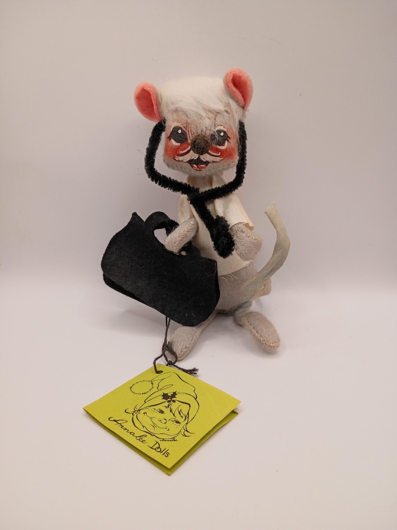 May include: A vintage Annalee Dolls mouse figurine, featuring a white head with pink ears, a black scarf, and a black bag. The mouse is holding a black bag and has a tag that says "Annalee Dolls".