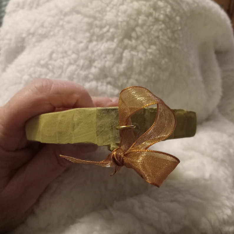 May include: A small, light green wooden object with a gold-coloured ribbon tied in a bow. The wood has a natural, unfinished look. A small gold ring is attached to the wood. The ribbon is a sheer, shimmering gold colour.