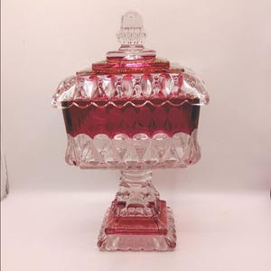 May include: A square glass candy dish featuring a deep red and clear finish. The piece includes a removable lid with a finial top, a faceted body with geometric patterns, and a square pedestal base. The design combines transparent glass with red.